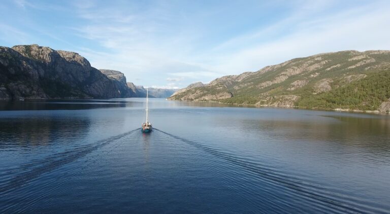 Hier zie je Noorwegen, de fjorden, in het midden vaart een zeilboot.